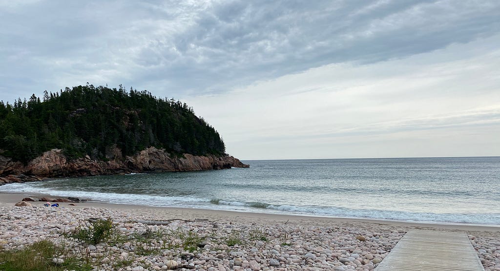 A pebbled beach with a boardwalk leading to the calm sea. On the left is a wooded headland.