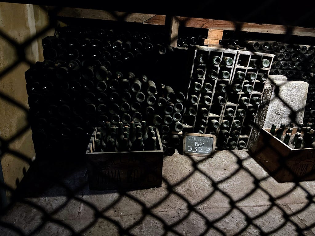 A dusty stash of 3000+ bottles of wine lying mostly on their sides and some in crates, with a small blackboard that reads “COLARES, 1969, 3323” with the later number clearly having been wiped out and amended multiple times (likely as wine is taken from the stash).