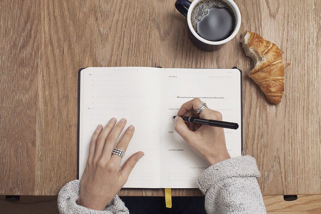A woman writing in a notebook wearing silver rings. There is a croissant and a cup of coffee next to her.
