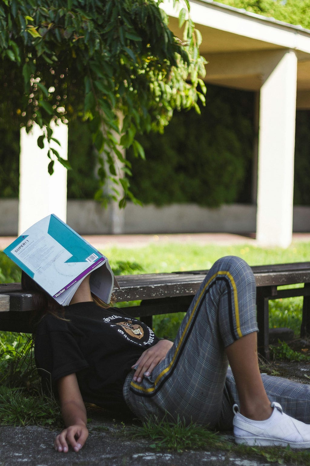 Girl resting her head on a bench with a blue book covering her face. She looks