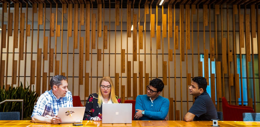 Four people, two with laptops, confer at a wood conference table in a corporate setting.