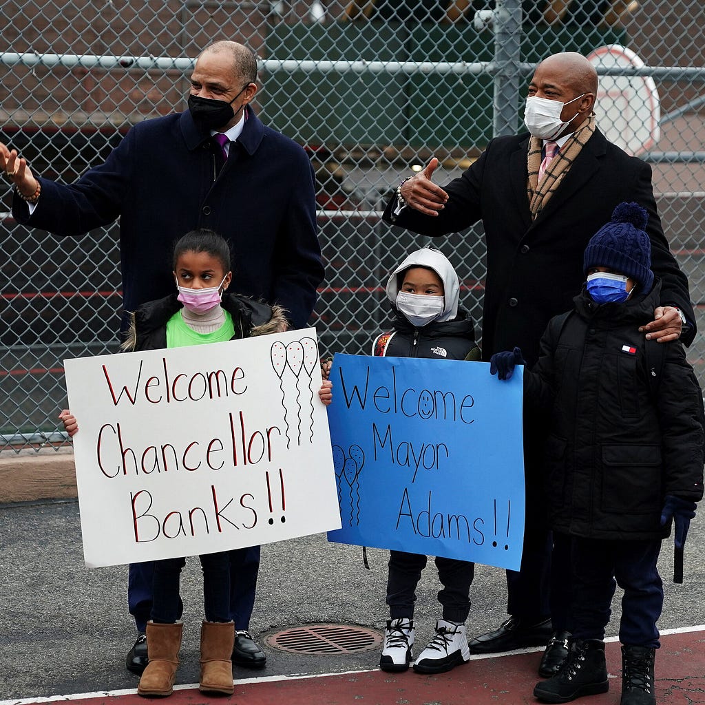 Chancellor David Banks and Mayor Eric Adams motion for people to join the photo op. Three children stand holding signs made by adults reading “Welcome Chancellor Banks” and “Welcome Mayor Adams!!” The child on the left side eyes while holding the sign.