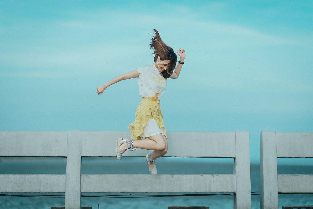 A young woman with a yellow skirt and white t-shirt jumping high against a blue sky and concrete fence.