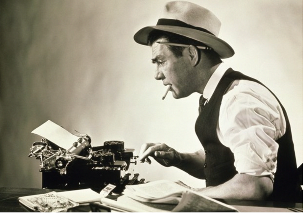 b&w photo of old fashioned jounralist at a typewriter wearing a hat smiking.
