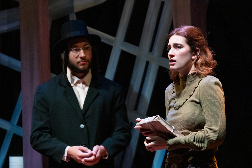 An actor wearing a black hat and coat with a beard and payes looks at an actress with short hair and a green sweater who is speaking while holding a book.