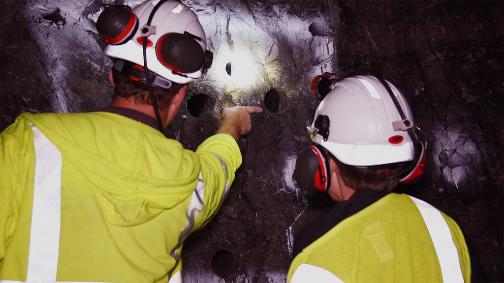 Workers examining drill holes in a mine wall.