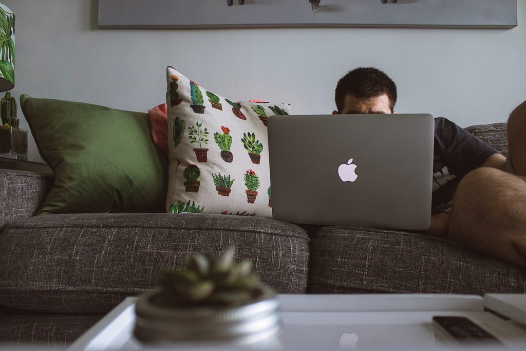 A couch is adorned with pillows and someone sits on the couch on a computer. Their head is obstructed by the laptop.