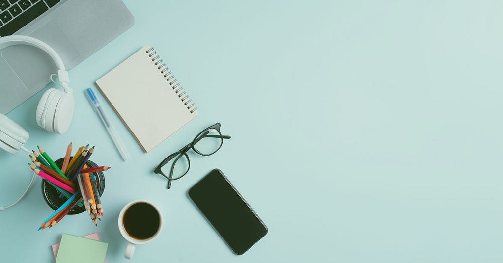 A notebook, smartphone, pencils, headphones, glasses, and a cup of coffee.