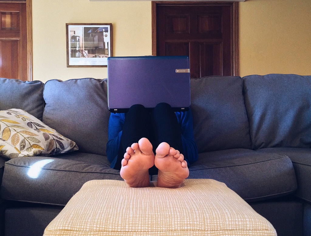 Someone sitting on the couch, feet on a stool, face hidden behind a laptop computer