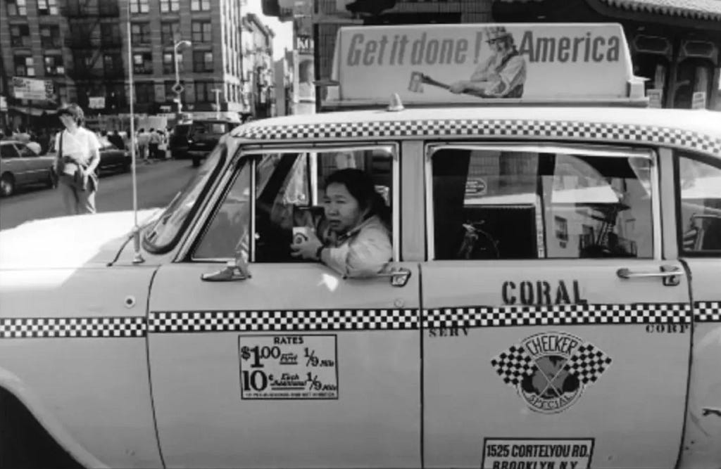 Chinese American woman taxi driver drinking coffee out of the side of a taxi that says “get it done america” on a sign