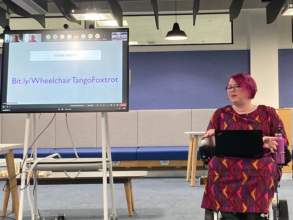 Claire in a bright dress, presenting from her chair. She has a laptop on her knees and is next to a screen with an online meeting open, showing a slide with the WTF blog url.