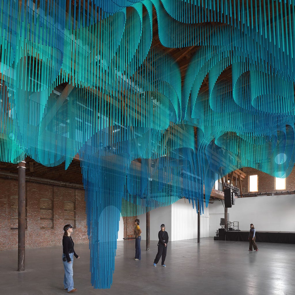 An industrial warehouse space, with a concrete floor and brick walls. People wander around, wearing black headsets. Above them is a rendering of a virtual entity: rows and rows of narrow slats of bright teal form a layered, undulating shape.