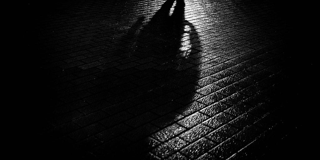 Long, threatening shadow of a man on wet, darkened pavement.