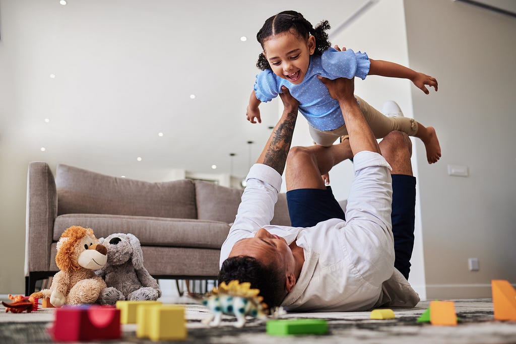 A happy father playing airplane with his daughter in a toy-filled living room, lifting her with his legs, bonding with love.