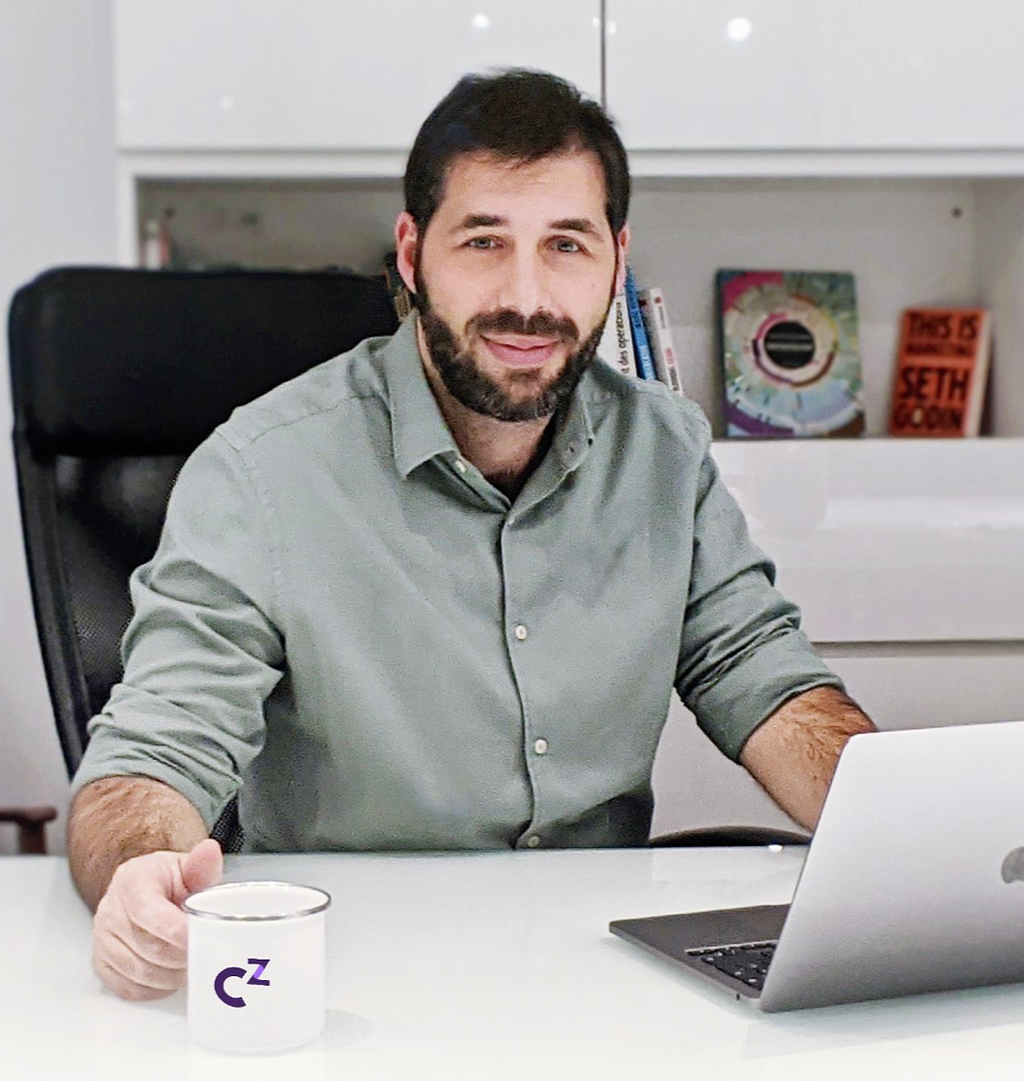 A man with dark hair and a beard sits at a work desk holding a cup of coffee.