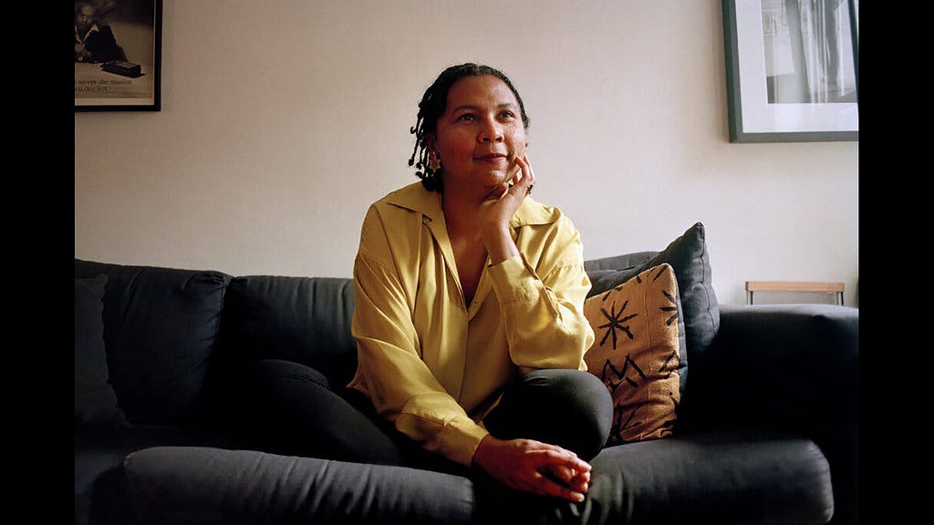 [Image ID: Feminist leader bell hooks on a couch with her chin resting on her hand, wearing a yellow shirt and looking thoughtful, per usual.]