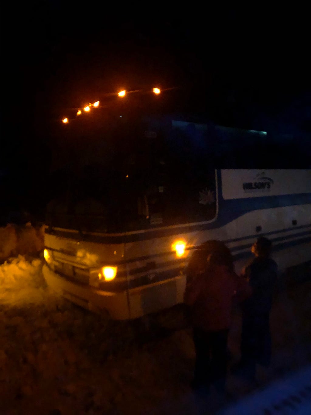 A picture of a coach bus by a snowy road at night. Two people stand by the side of the bus looking at it.