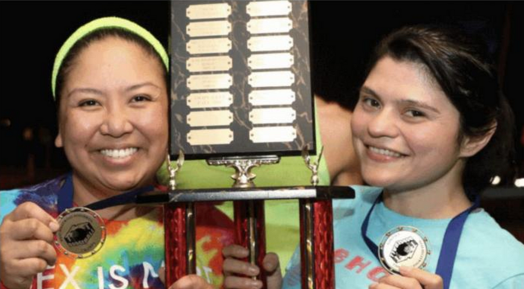 A close-up image of two members from WeHo Dodgeball smiling proudly, both showing off the medals around their necks while holding on to the large trophy between them.