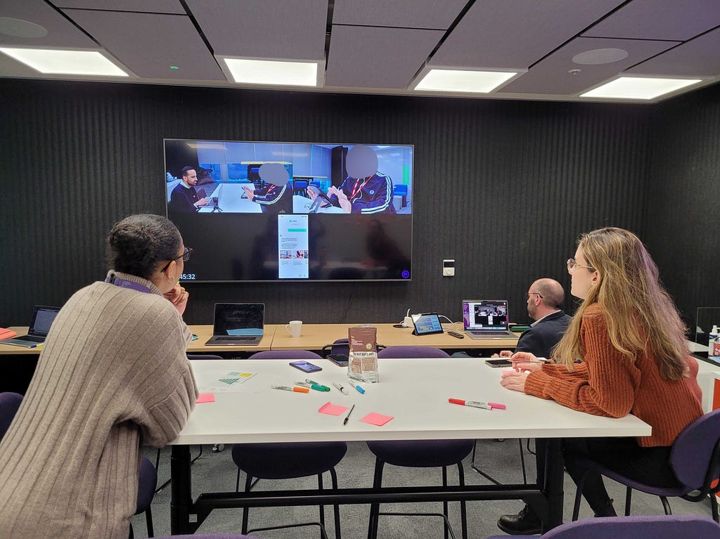 In an office, 3 user researchers are watching a live video feed attached to the wall. On the feed is a split screen, showing 2 users interacting with new digital products and describing their experiences (their faces are blurred) The researchers have pens and post-it’s in front of them as they take notes.