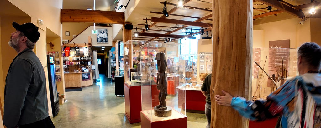 Interior of the Duwamish Longhouse museum and gift shop, with cultural exhibits, wood beams, and visitors exploring the space. A carved wooden statue stands in a glass case at center.