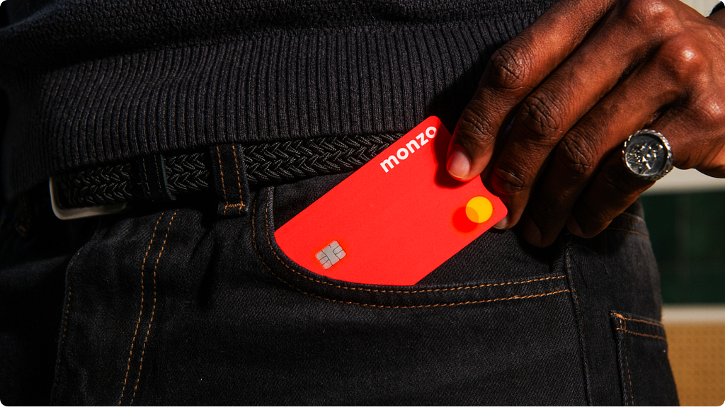 A bright coral Monzo card half-tucked into the back pocket of dark indigo jeans, with a hand drawing it out.