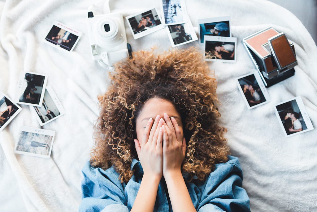 A woman laying on a white blanket, her face covered by her hands. Surrounding her like a rainbow over her head are a series of instant photos.