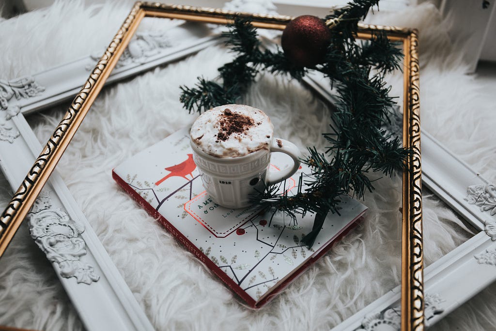 Empty frames with wintergreen garland and ornament, in center is cup of hot chocolate on top of a book called Merry Christmas