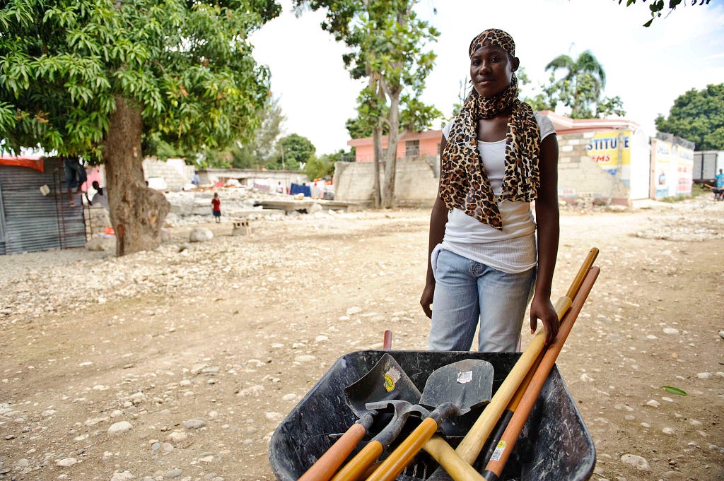 A woman pushes a wheelbarrow filled with spades and other less visible tools, along a dirt road. She is wearing a leapord-skin print headscarf, a white tank top and jeans.