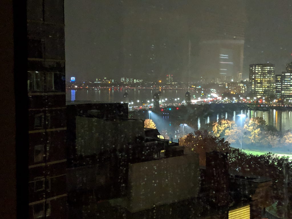 A view through a window of the Longfellow Bridge lit up on a rainy night. In the far distance is a tiny neon Citgo sign.