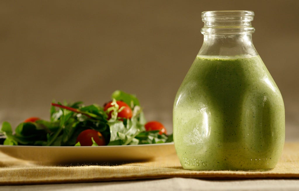 A short glass bottle of green dressing positioned between the camera and a plate heaped with salad.