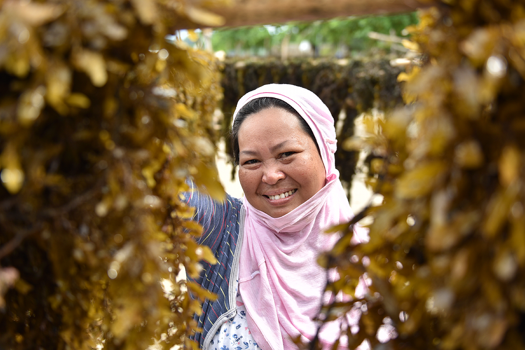 A smiling woman looks through an opening in a harvest of seagrass.