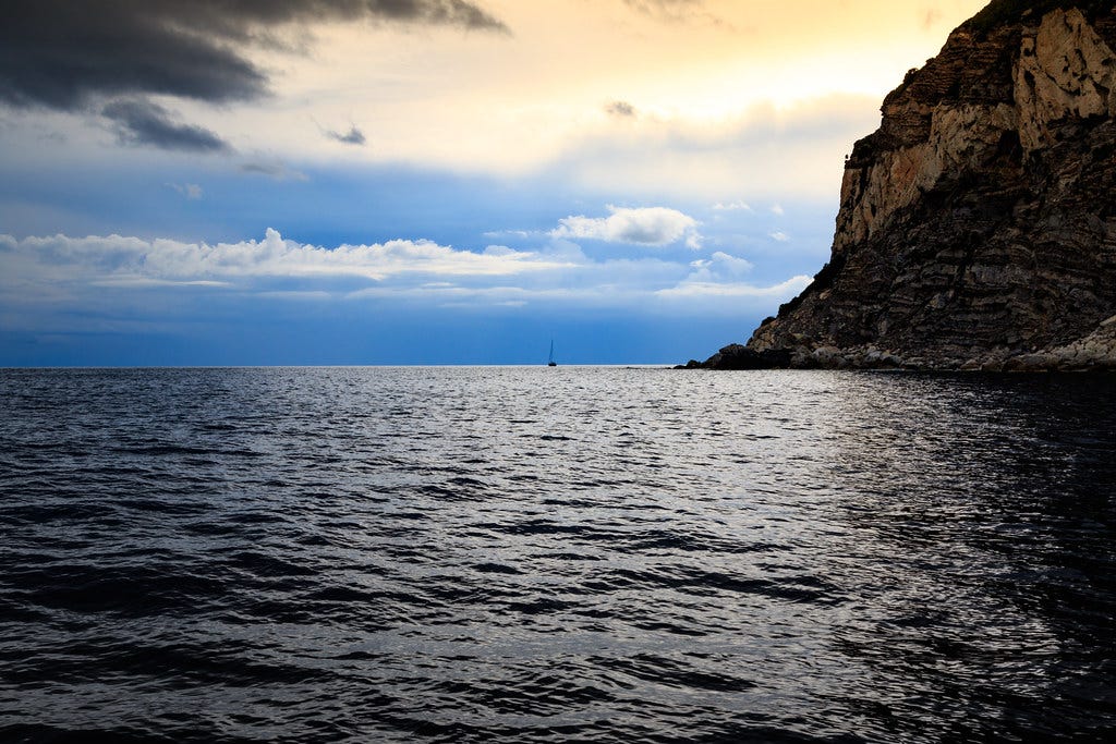 A single ship in the distance near the horizon, water in foreground, blue/pink sky in the background, cliffs on the right.