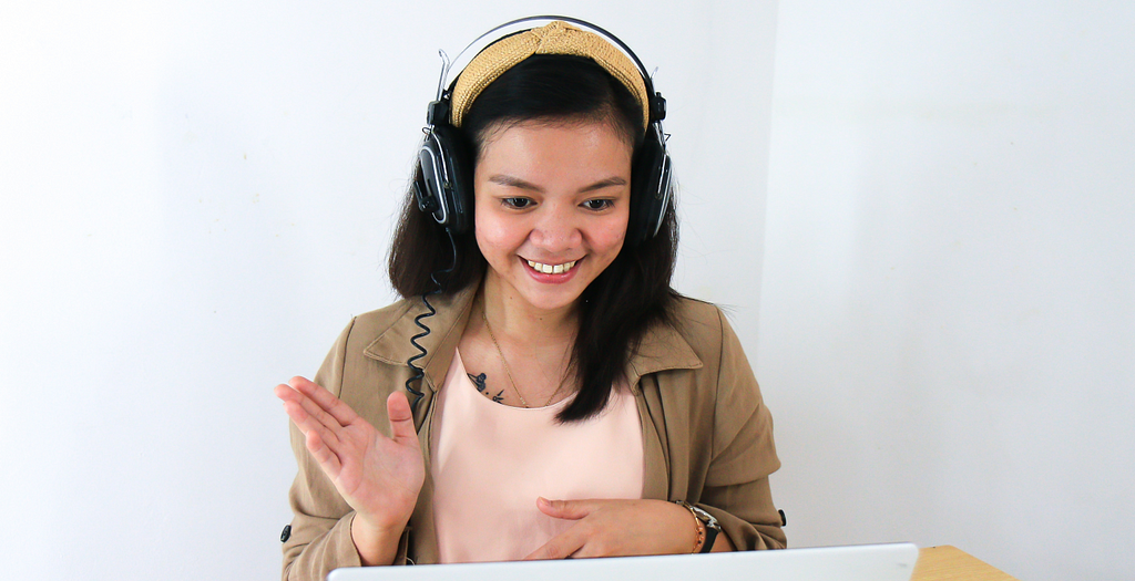 A woman wearing headphones, smiling and waving at a laptop screen
