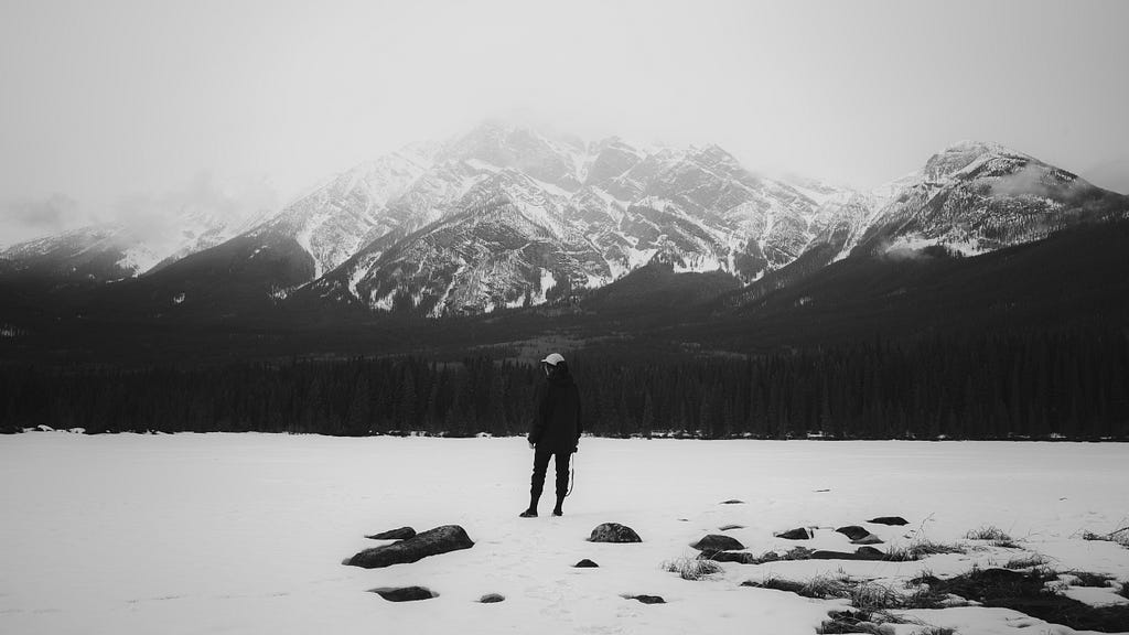 A woman stands on a frozen lake, a towering mountain looms in the clouds beyond her.