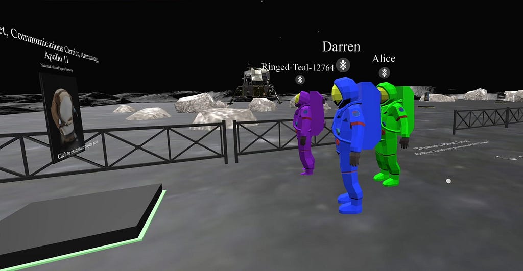 Three players’ avatars in space suits that are bright shades of purple, blue, and green, stand in the moonscape in front of a floating image of a “communications device” from Apollo 11. Each player’s name floats above their avatar’s head. A black fence, scattered moon boulders, and a moon lander are visible in the background.