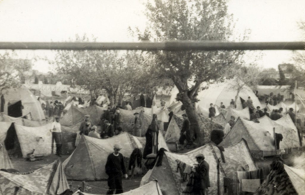 A black-and-white image of a group of about one dozen white tents and a tree in the middle. Refugees from the Pentcho ship walk amid the tents.