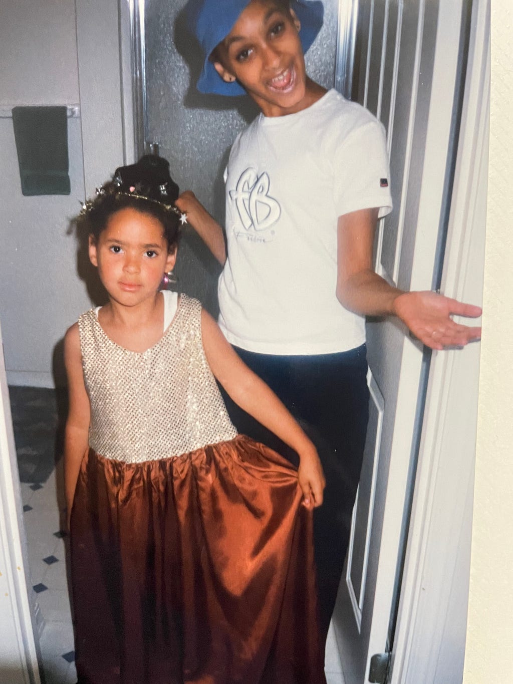 Me probably around age 7 and my older sister around age 12, I’m wearing a dress with a light gold sparkly top and a satiny brown skirt with all manner of ribbon and metallic glitz in my done up hair. Whitney is dramatically presenting me with her hands out sporting a white FUBU shirt and a blue bucket hat.