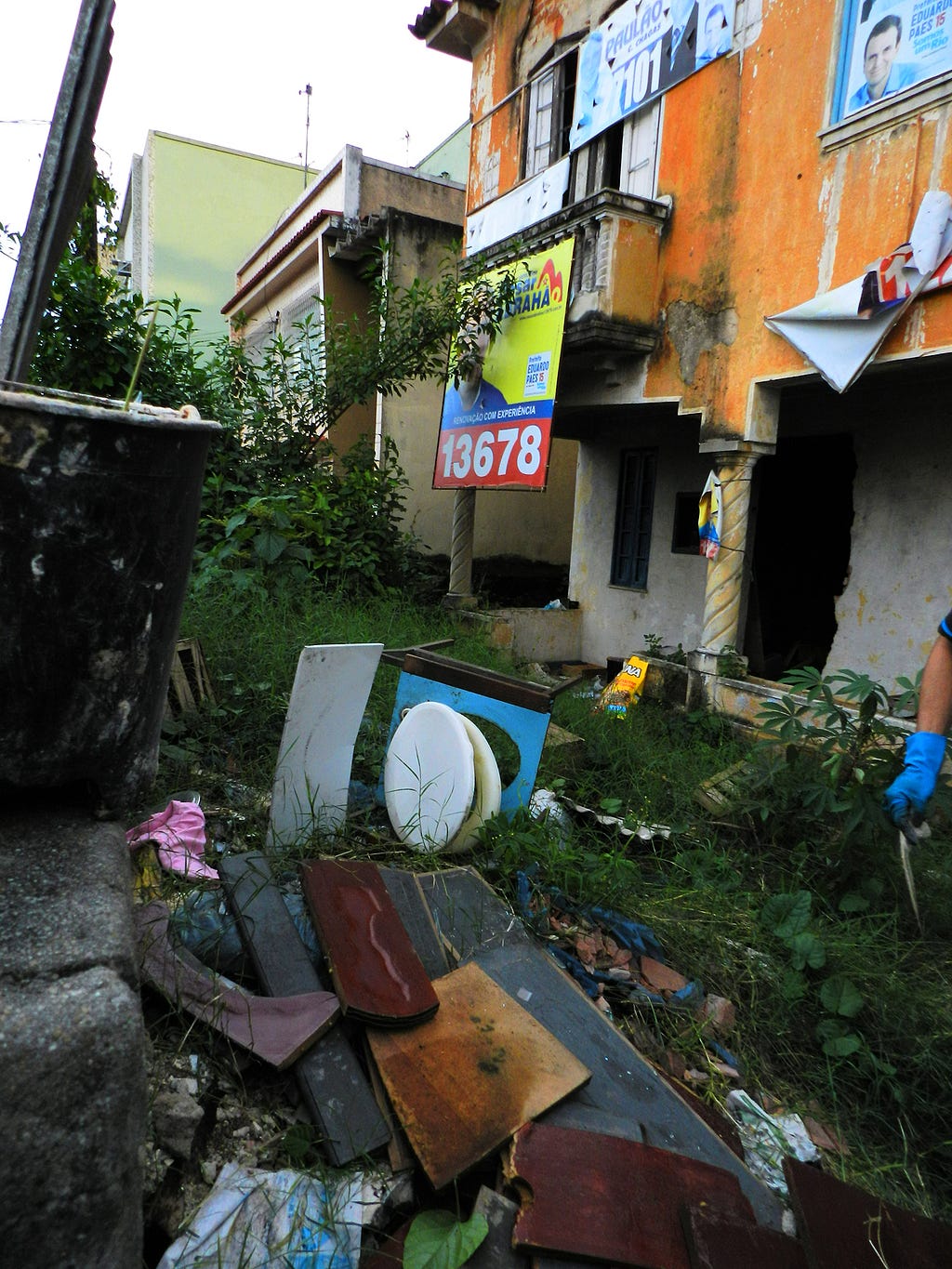 Photo of the house front yard with tall grass and variety of dispoased objects such as toilet seat, clothes and furniture.