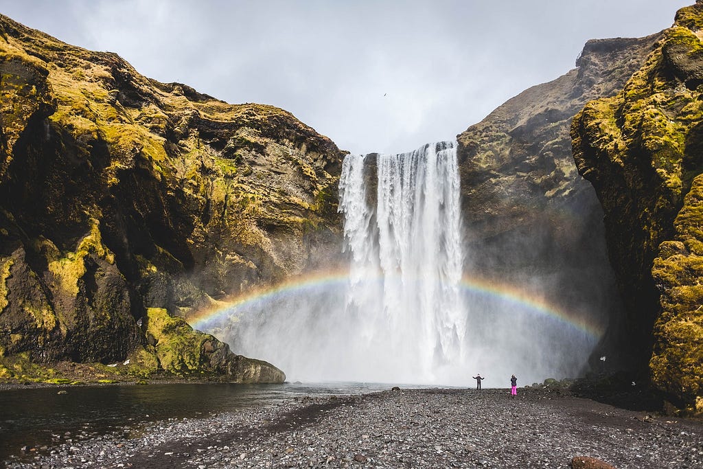 Stream of water falling downward from a cliff creating a waterfall with a rainbow behind it