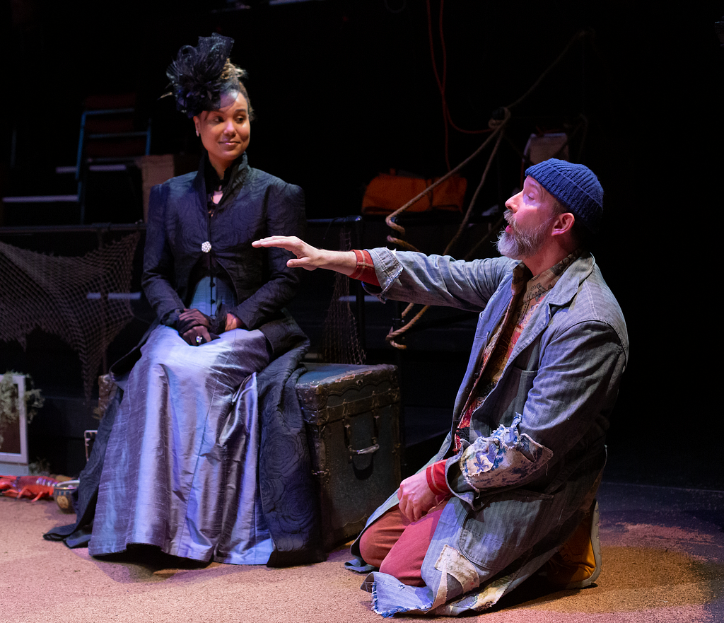 A woman in 19th century-inspired mourning clothes sits and smiles at a man kneeling and holding out a hand to her in a denim jacket and a knit hat.