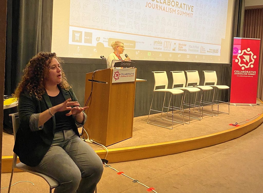 An ASL interpreter signs at the front of an auditorium as Stefanie Murray stands at a podium on stage and gives the opening remarks on day one of the 2022 Collaborative Journalism Summit.