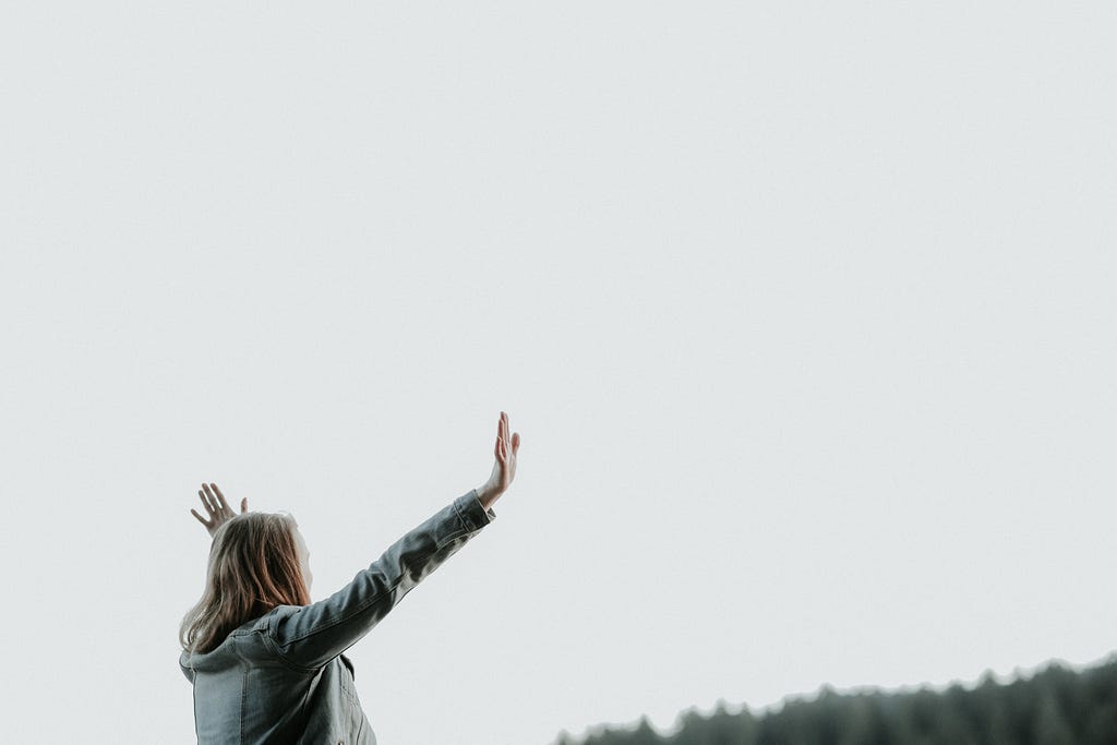 A woman stands on a hillside, the sky is grey but she has her hands raised in worship to God. We see her as a side-on view and cannot see her face, but only her pose of focus towards God, surrounded by His creation.