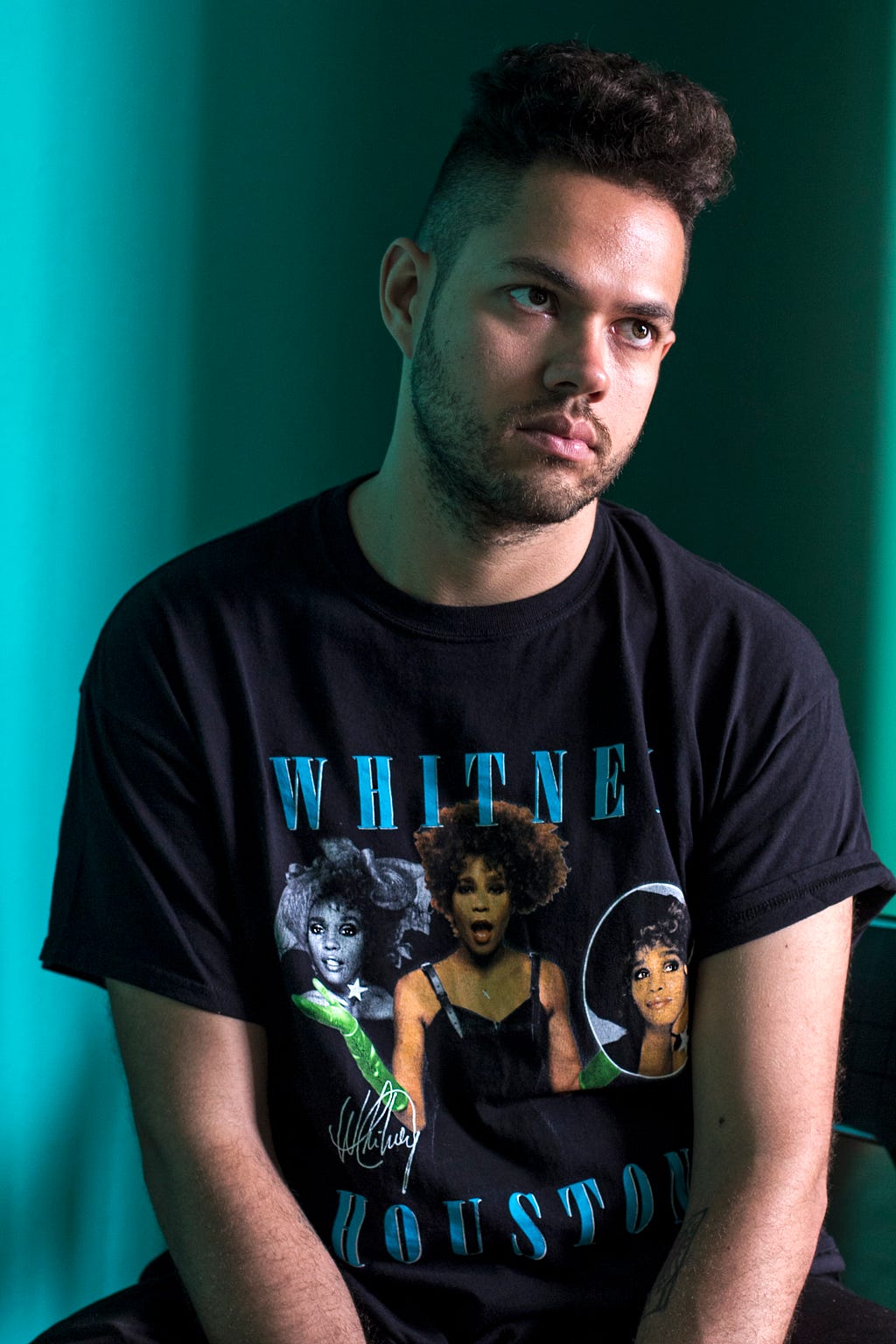 Foto do Gustavo Abreu. Estávestindo camiseta preta, tem barba e está com um fundo escuro atrás