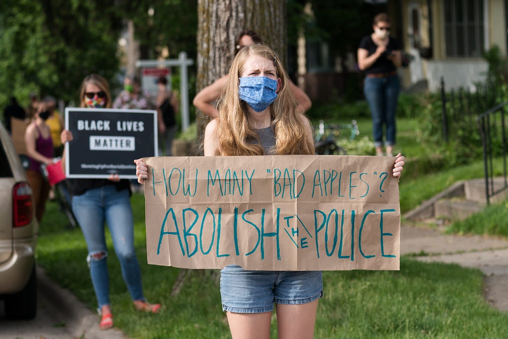 A masked woman holds a sign reading “HOW MANY ‘BAD APPLES’? ABOLISH THE POLICE”. In the background, another woman holds a sign reading “BLACK LIVES MATTER”.