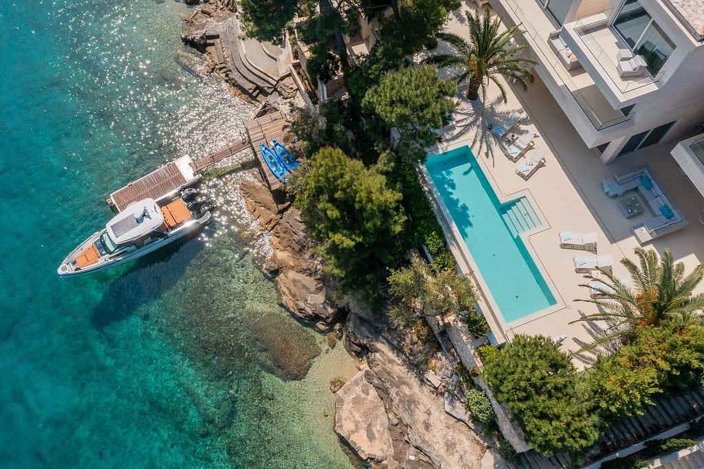 Aerial view of a luxurious coastal villa with an infinity pool, spacious sun deck, and modern outdoor furniture. The villa is nestled among lush greenery and tall palm trees, offering a serene and private atmosphere. A wooden dock extends into the clear turquoise waters, where a sleek yacht is moored alongside several kayaks. The rocky shoreline adds natural beauty to the setting, with steps leading down to the water. The villa’s balconies and terraces provide stunning views of the sparkling sea
