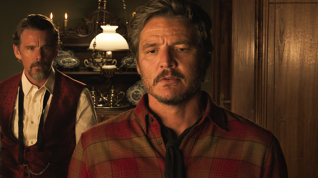 Silva (Pedro Pascal) is standing in a parlour room facing the camera. To the left, behind him, Jake (Ethan Hawke) looks longingly over Silva’s shoulder.