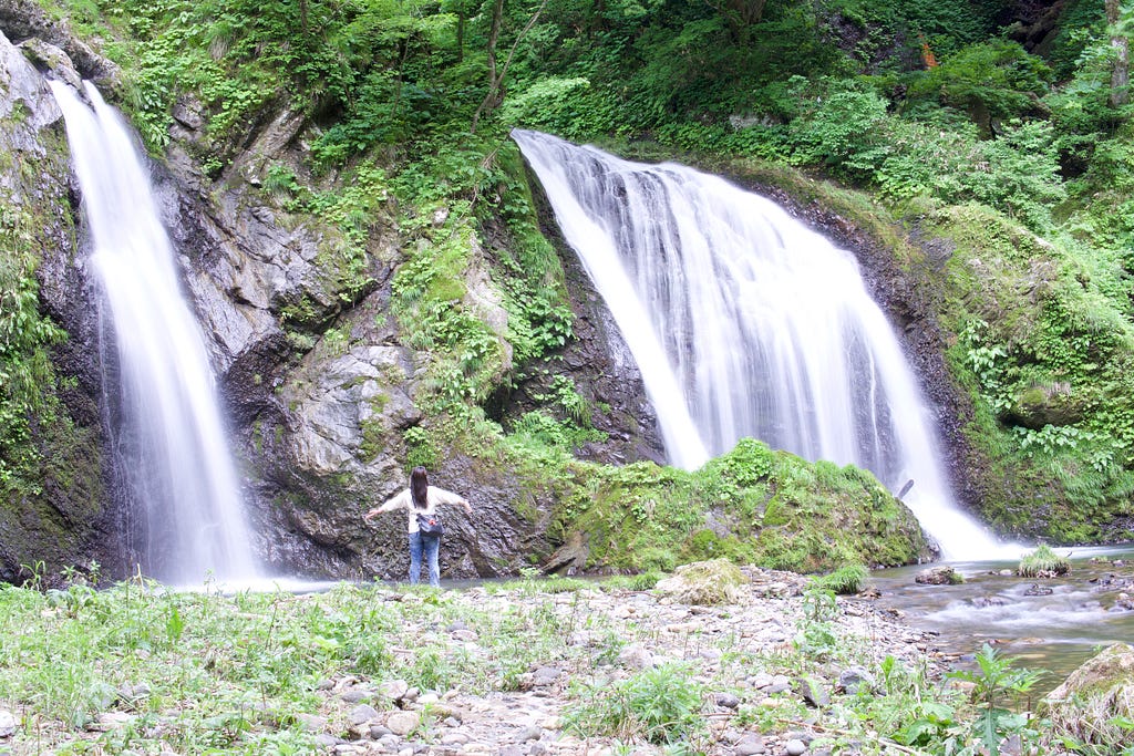 A person stands arms stretched wide in front of Juninotaki Falls at the base of Mt. Kyogakura, one of the 100 Famous Mountains of Yamagata located in Sakata City, in the Tohoku region of North Japan.