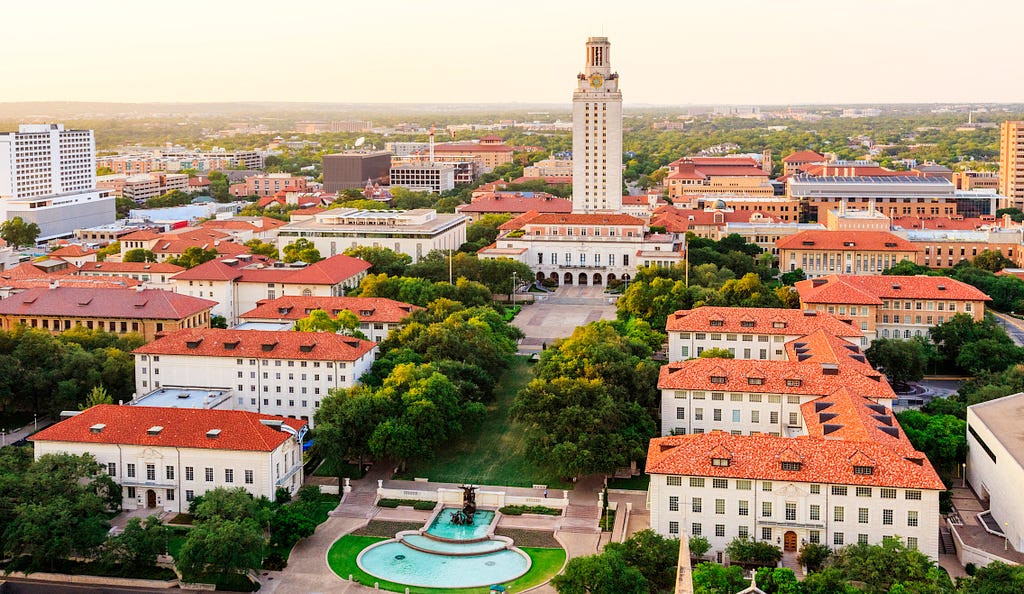 Universidade do Texas em Austin vista de cima, há diversas árvores ao redor do campus e diversos edifícios com telhados marrom-claros. Há uma fonte em destaque no centro da foto.