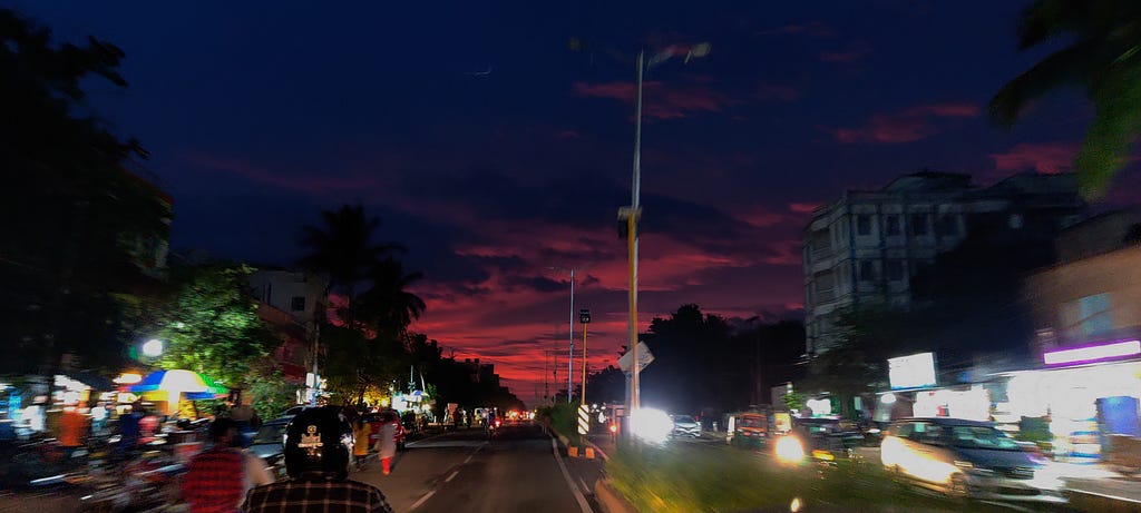 a not-so-usual evening sky, just a few minutes after dusk. The sky is shades of purple and pink with more pink towards the horizon. It is a mildly busy two-way road, a darker setting being lit up by the headlights of the vehicles.