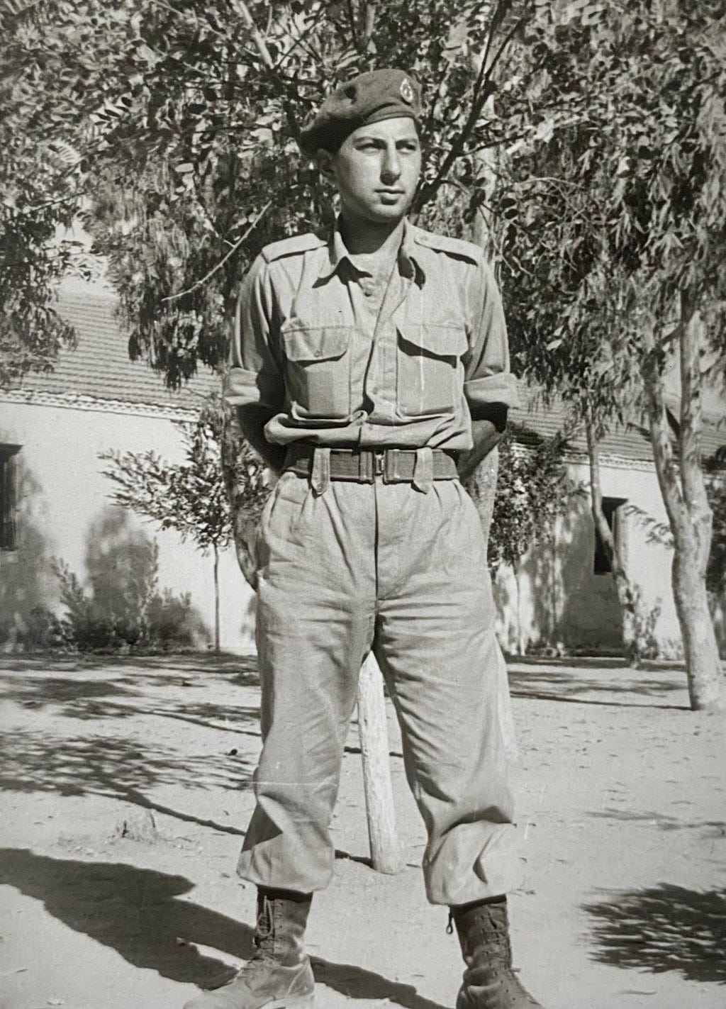 A dark haired man in a military uniform standing with his legs spread, arms behind his back.